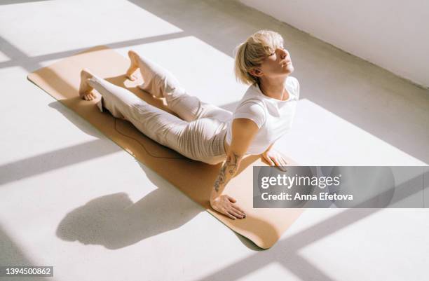 beautiful authentic woman with tattoos and short blond hair is doing yoga in upward facing dog position on yoga mat in front of a window. she is wearing a light-colored casual clothing. concept of relaxation exercises - upright position stock pictures, royalty-free photos & images