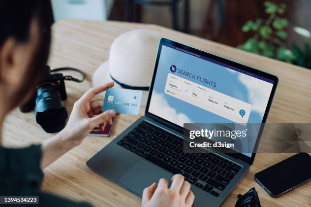 over the shoulder view of young asian woman shopping online for flight tickets on airline website with laptop, entering credit card details to make mobile payment at home. camera and passport on the table. travel planning. booking a holiday online - ordering stock-fotos und bilder