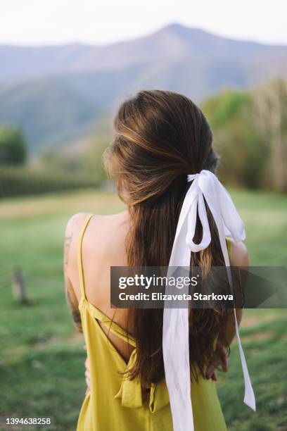 a large portrait of a fashionable hairstyle with a white bow of a young woman with tattoos in a dress walking in nature on vacation traveling against the background of mountains and trees standing with her back. - hair bow stock pictures, royalty-free photos & images
