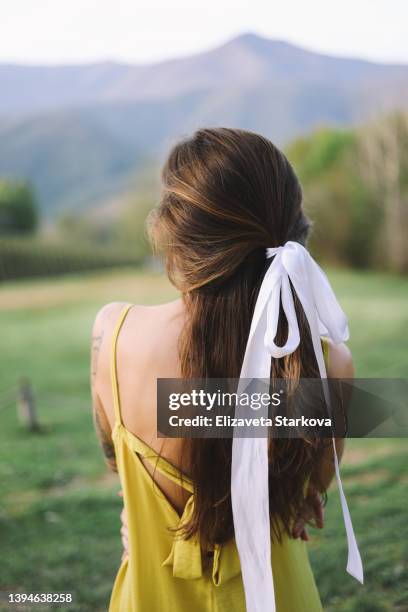 a large portrait of a fashionable hairstyle with a white bow of a young woman with tattoos in a dress walking in nature on vacation traveling against the background of mountains and trees standing with her back. - hair bow imagens e fotografias de stock