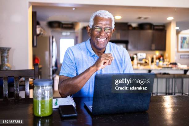 senior black man working at laptop at dining table - stralende lach stockfoto's en -beelden