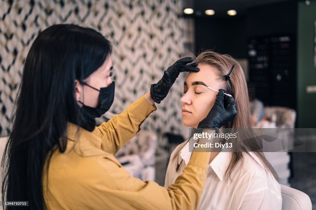 A master cosmetologist paints the eyebrows of a client in the salon close-up. eyebrow dyeing procedure, girl wipes paint from her eyebrows.