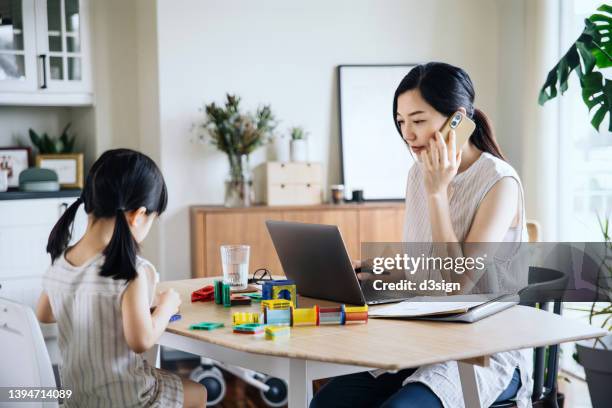 concentrated young asian mother working from home on laptop and talking on the phone while little daughter is playing with toy blocks on the desk. home office and business concept. working mom managing work life and home life - working mum stock pictures, royalty-free photos & images