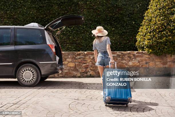 a young woman pulling a suitcase to load in an opened trunk car on summer holidays. travel concept - dragging fotografías e imágenes de stock