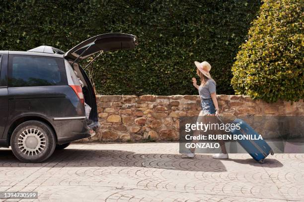 a young woman pulling a suitcase to load in an opened trunk car on summer holidays. travel concept - white van profile stock pictures, royalty-free photos & images