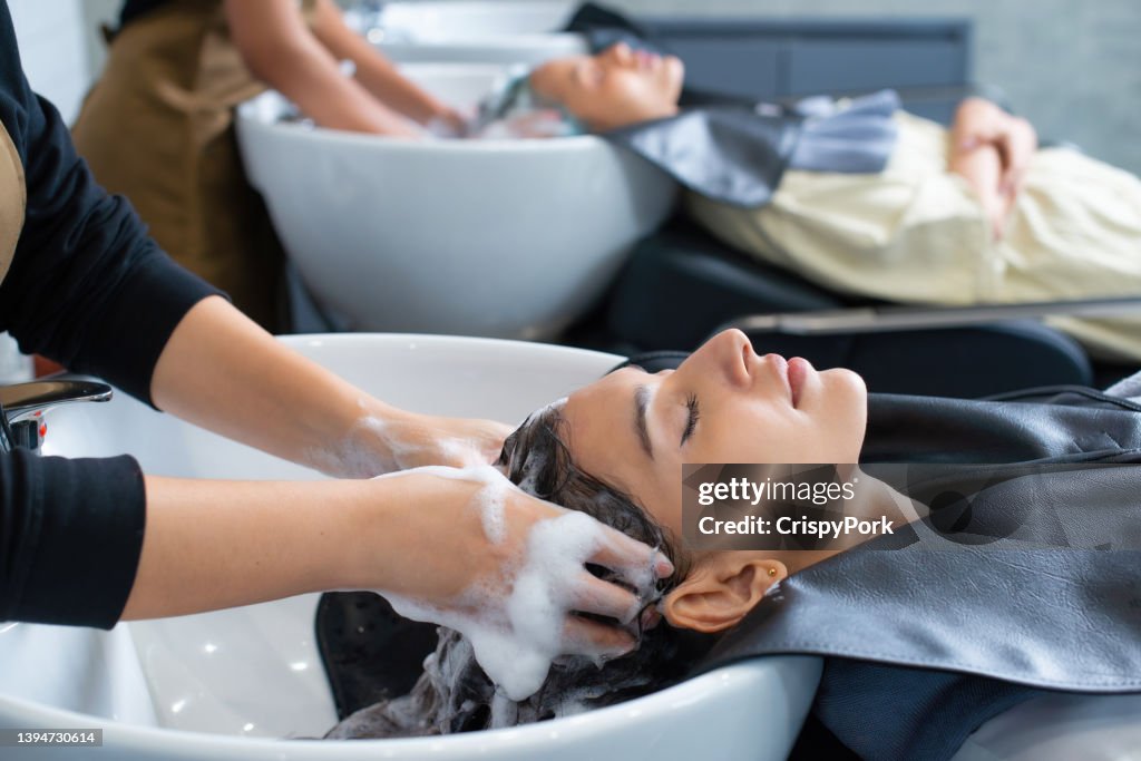 Young woman in hairdresser salon during hair wash after haircut. Woman applying shampoo and massaging hair of a customer. Female having her hair washed in a hairdressing salon.