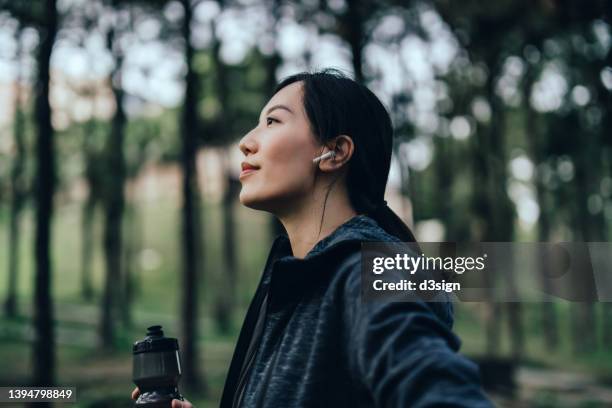 confident and determined young asian sports woman with wireless earphones holding a water bottle, getting ready to work out outdoors in nature park. health and fitness training routine. healthy living lifestyle - best female performance stock pictures, royalty-free photos & images