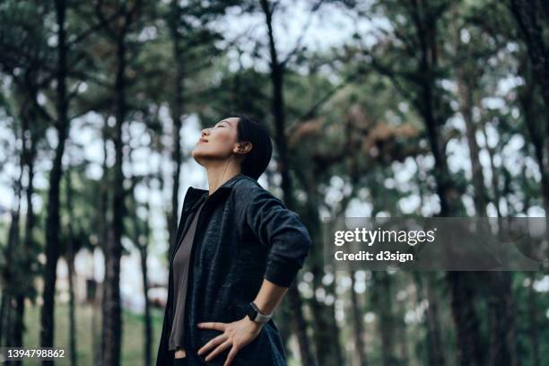 relaxed young asian sports woman with eyes closed breathing fresh air in nature park with trees in background. connection with nature. freedom in nature - anticipation sport stock pictures, royalty-free photos & images