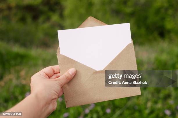 envelope with blank white paper card for copy space held in hand against green leaves and flowers floral background - holding card stock pictures, royalty-free photos & images