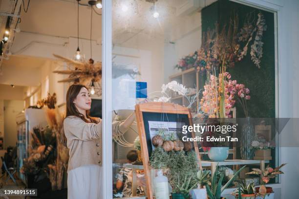 asiatische chinesische floristin geschäftsinhaberin wiedereröffnung für geschäfte und öffnet schild am glasfenster vor ihrem blumenladen - open sign on door stock-fotos und bilder