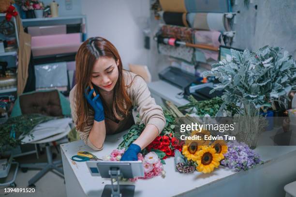 asian chine female florist, small business flower shop owner, talking on smartphone while working on digital tablet in workplace. checking stocks, taking customer orders, selling products online. daily routine of running a small business with technology - chine business stock pictures, royalty-free photos & images