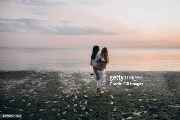 mother carrying daughter by the sea at dusk - wide stock-fotos und bilder
