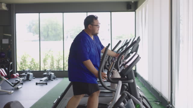 4K Side view of overweight man jogging on eliptical machine at indoor gym.