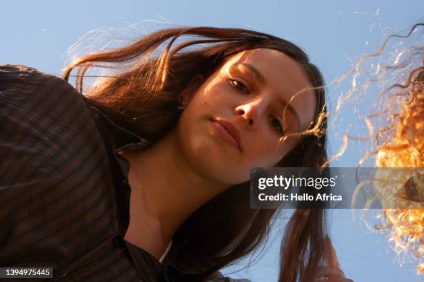 a beautiful teenage girl with long straight loose brown hair looks down at camera with a friend's shock of curly hair sneaking into the frame with a backdrop of clear blue sky - offbeat stock pictures, royalty-free photos & images