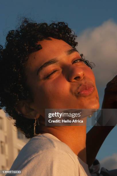 a beautiful young woman with short curly hair stands smiling looking at camera with her head at an angle & whilst both winking with an eye closed and pouting her lips as if to blow the viewer a kiss - blowing kiss stock pictures, royalty-free photos & images