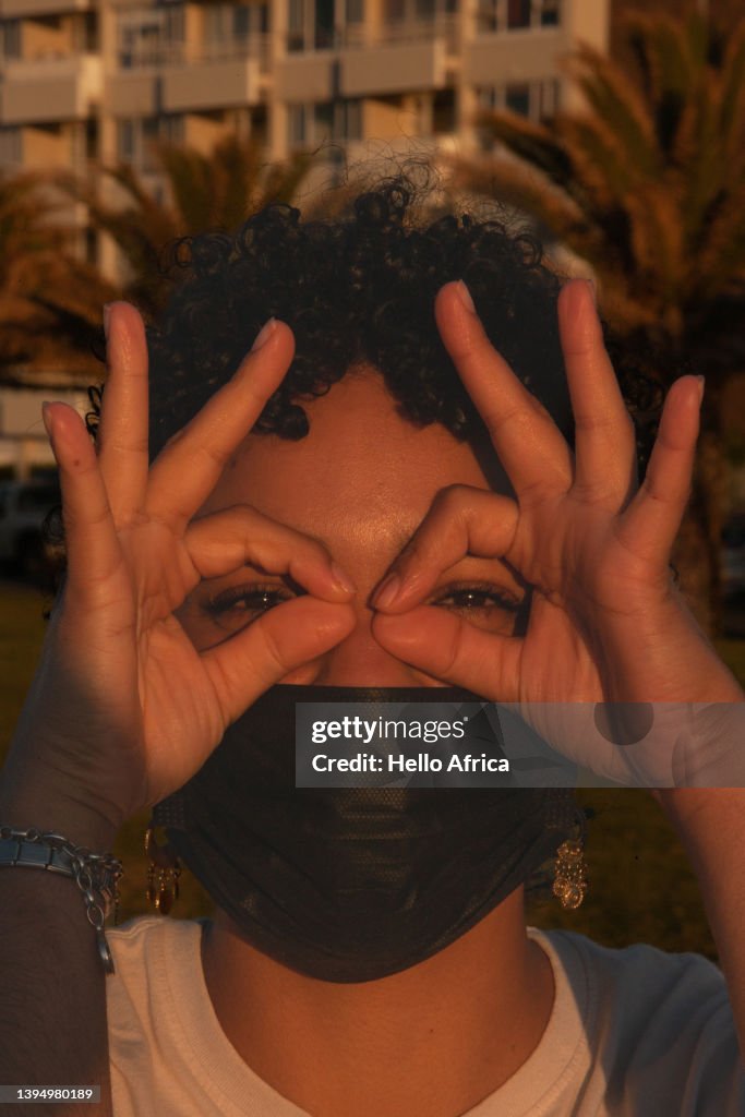 Vertical orientation portrait of a beautiful young woman with short black curly hair looks with her eyes through the eyes of the perfect/ perfection gestures she’s making with her fingers & hands