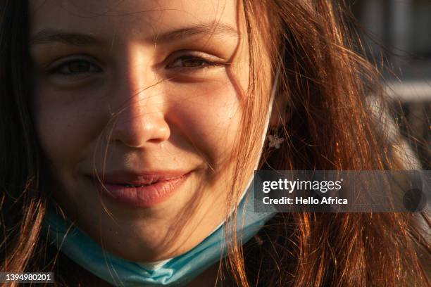 landscape portrait of a beautiful widely smiling teenage girl, with long flowing brown hair & with her blue & white disposable surgical face mask tucked under her chin giving a sense of freedom following stricter quarantine times - giving a girl head stock pictures, royalty-free photos & images