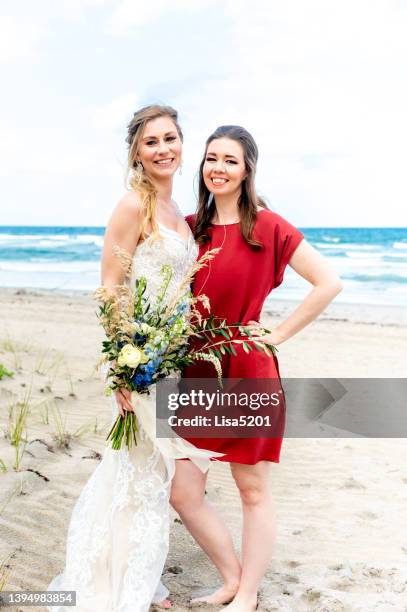 two beautiful women on the beach, one woman wears a wedding dress and holds a bouquet of flowers, destination wedding or elopement - bridesmaid stock pictures, royalty-free photos & images