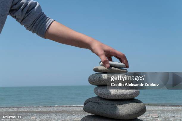 a man or a teenager makes, builds and creates a pyramid of sea stones or large pebbles on the seashore, on the beach, against the horizon. the concept of harmony and balance in the soul. a man is holding a stone in his hands, trying to find a balance. - georgian man stock pictures, royalty-free photos & images