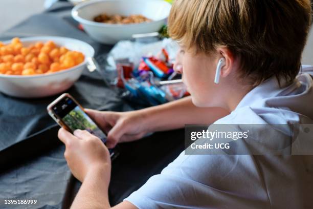 Preteen boy concentrating on smartphone with ear buds and bowls of snacks.