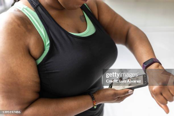 plus size woman comparing the data on her smartwatch and phone at fitness studio - exercise watch stock pictures, royalty-free photos & images