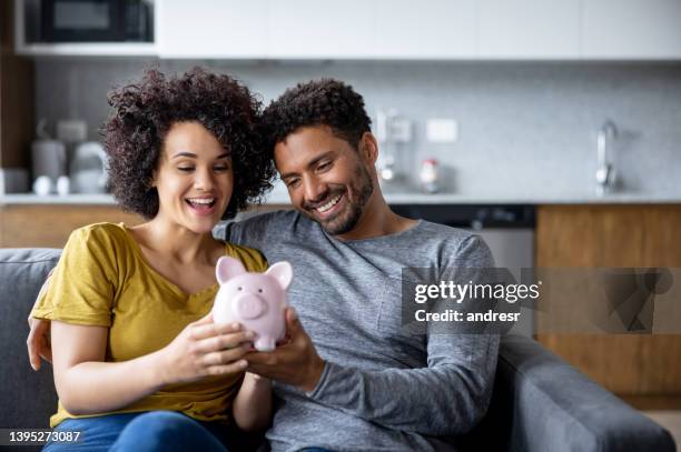 glückliches paar zu hause, das mit seinen ersparnissen ein sparschwein hält - couple saving piggy bank stock-fotos und bilder