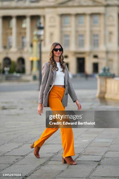 Amanda Derhy wears black sunglasses, a white cropped t-shirt, a gold chain pendant necklace, a brown and beige houndstooths print pattern long blazer...