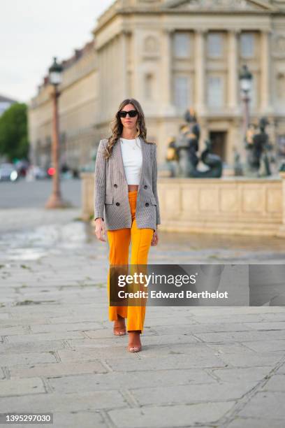 Amanda Derhy wears black sunglasses, a white cropped t-shirt, a gold chain pendant necklace, a brown and beige houndstooths print pattern long blazer...