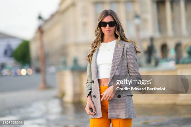Amanda Derhy wears black sunglasses, a white cropped t-shirt, a gold chain pendant necklace, a brown and beige houndstooths print pattern long blazer...