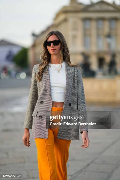 Amanda Derhy wears black sunglasses, a white cropped t-shirt, a gold chain pendant necklace, a brown and beige houndstooths print pattern long blazer...