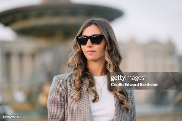 Amanda Derhy wears black sunglasses, a white cropped t-shirt, a gold chain pendant necklace, a brown and beige houndstooths print pattern long blazer...