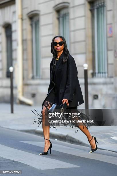 Emilie Joseph wears black sunglasses, rhinestone earrings, a gold large chain necklace, a black embroidered short dress with long fringed details, a...