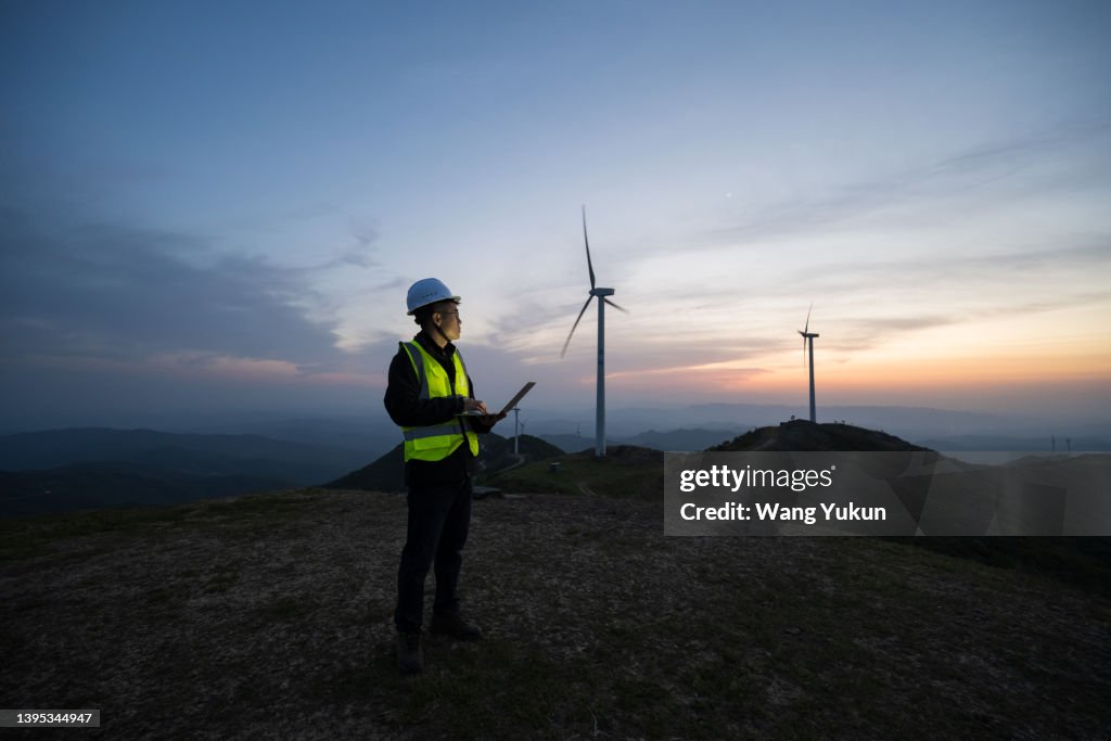 An Asian male electrical engineer is standing at a wind power plant with a laptop in the evening