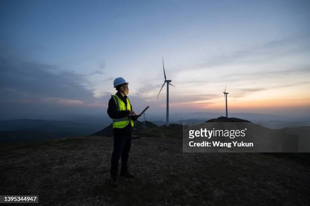 an asian male electrical engineer is standing at a wind power plant with a laptop in the evening - dusk stock photos et images de collection