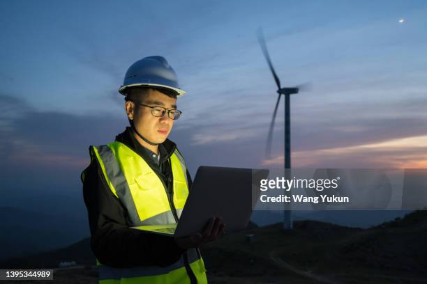 an asian male electrical engineer is standing at a wind power plant with a laptop in the evening - confidence male landscape stock-fotos und bilder