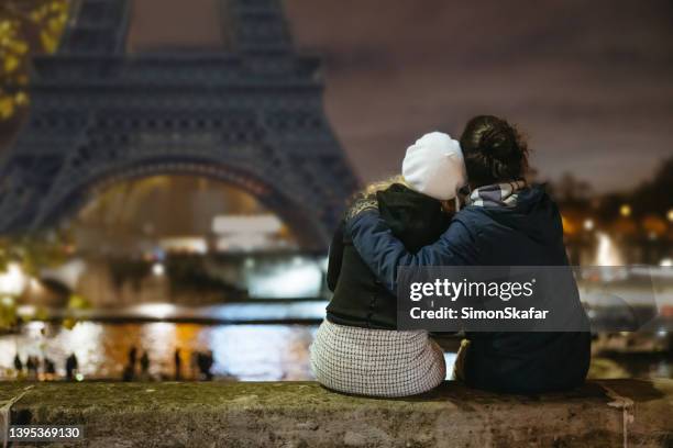 romantic couple sitting on a wall at seine river, looking at eiffel tower, paris in the evening - paris night stock pictures, royalty-free photos & images