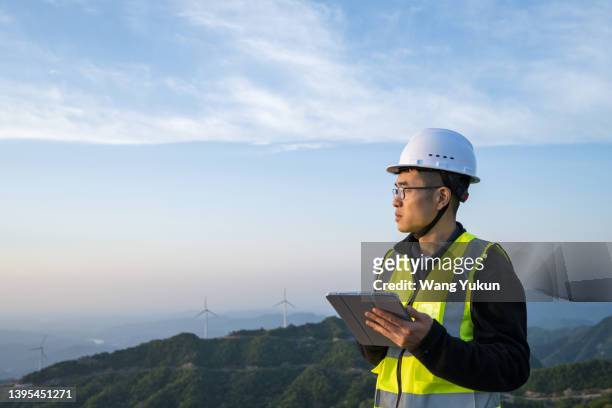 an east asian male power engineer holds a tablet computer and stands in the profile of a wind power plant - looks right stockfoto's en -beelden