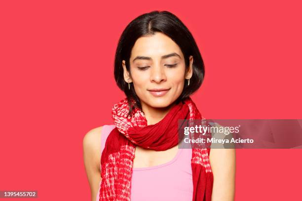 portrait of a fashionable indian millennial woman happily wearing a red scarf, a pink tank top and turquoise earrings, stands in front of a red backdrop in a studio setting. - red scarf stock-fotos und bilder