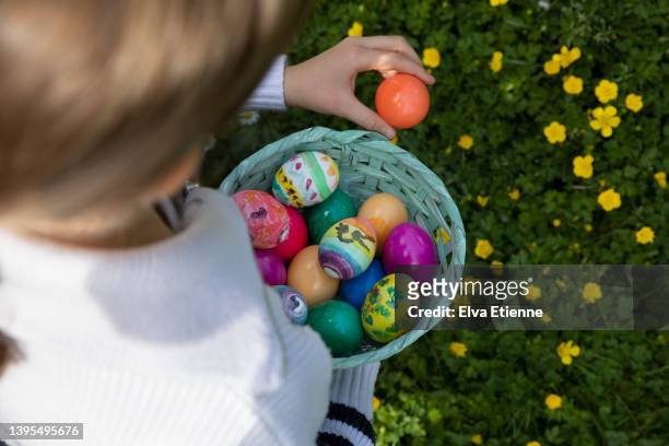 high angle view of a child collecting multicoloured painted easter eggs in a basket made of woven cardboard in a back yard. - eggs basket stock pictures, royalty-free photos & images