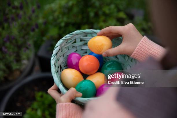 over the shoulder view of a child holding an easter basket made of woven cardboard and filled with multicoloured dyed hens eggs in a back yard. - easter egg hunt stock pictures, royalty-free photos & images