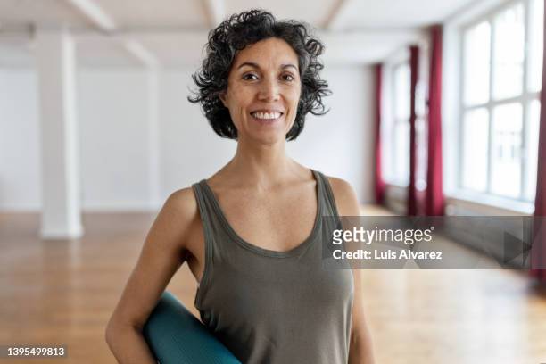 portrait of a happy mature woman with yoga mat standing in gym studio - yoga pose photos et images de collection