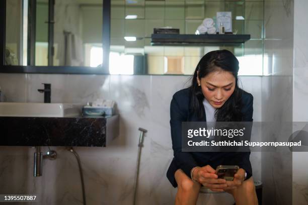 asian happy woman using smart phone while sitting on toilet seat in bathroom. - woman toilet stock pictures, royalty-free photos & images