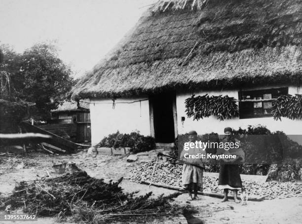 Barefoot women, one holding a sickle, stand next to crops, including corn hanged to dry, near a traditional barn in Ukraine, Soviet Union, circa 1920.