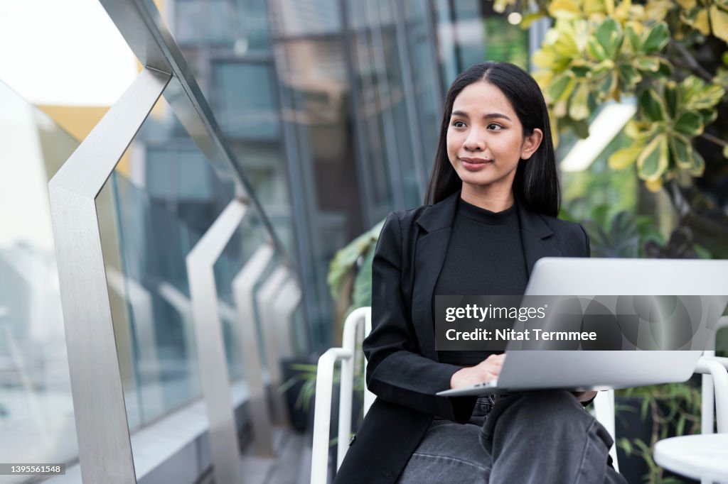 Working Remotely for The New Generation Of Creative Entrepreneurs. Young Asian Business woman working on laptop while looking away, sitting at bar rooftop hotel.