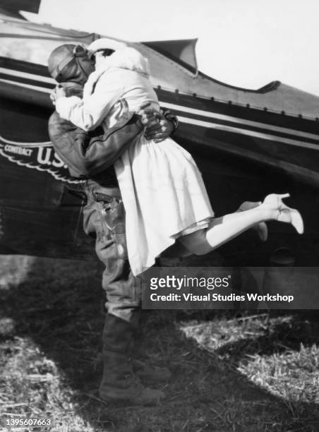 America pilot Alva R DeGarmo , in his Western Air Express flight suit) lifts his wife for a kiss as he stands beside a plane, Los Angeles,...