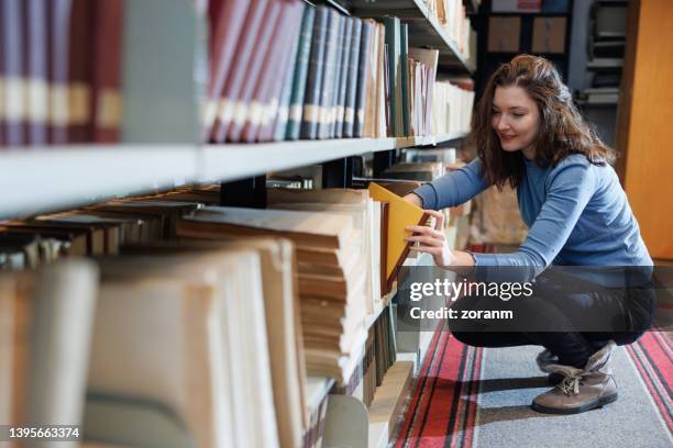 female post-grad crouching by the shelves in university library, searching for material - librarian stock pictures, royalty-free photos & images