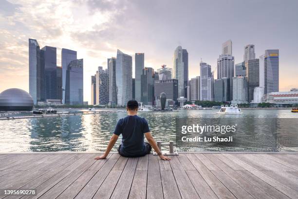 the young man sat on the dock with urban skyline and skyscrapers in marina bay singapore. - singapore tourist stock pictures, royalty-free photos & images