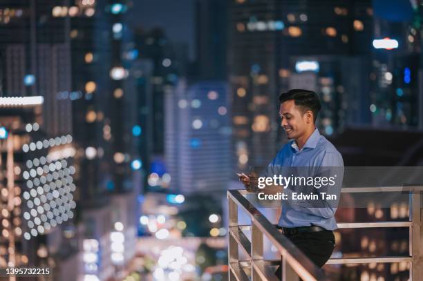 asian indian white collar man outside of office building taking a break using phone texting with city skyscraper - man on top of building stock pictures, royalty-free photos & images