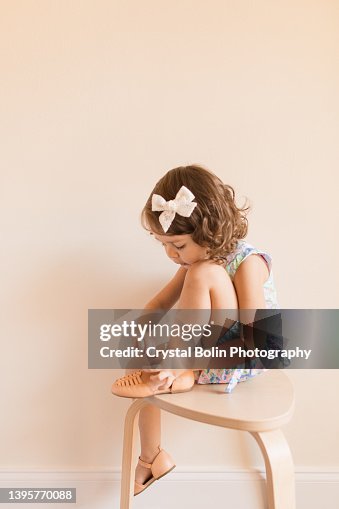 3-Year-Old Toddler Girl Dressed in a Pastel Shift Dress & White Bow, Sitting on a Chair Putting Her Shoes On 3-Year-Old Toddler Girl Dressed in a Pastel Shift Dress & White Bow, Sitting on a Chair Putting Her Shoes On