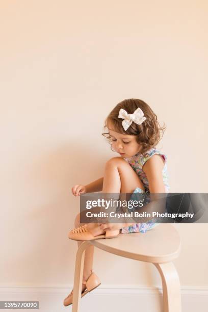 3-year-old toddler girl dressed in a pastel shift dress & white bow, sitting on a chair putting her shoes on - beige shoe stock pictures, royalty-free photos & images